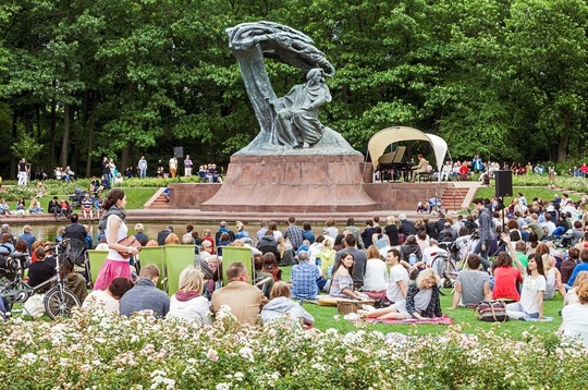 Concerto di pianoforte nel parco, persone sedute tra i fiori sull'erba, statua di Chopin